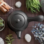 Composición zen con tetera de cerámica negra, canela, lavanda, piedras y plantas sobre esterilla de bambú